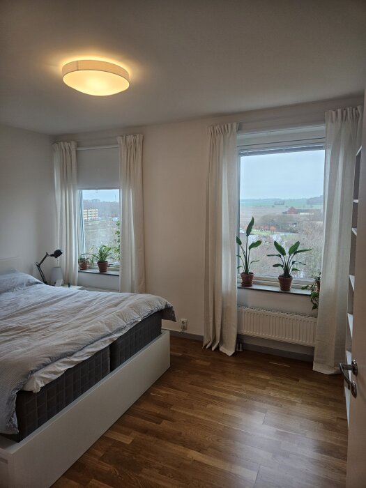 Sovrum med dubbelsäng, träparkettgolv, vita väggar och två fönster med växter på fönsterbrädorna. Taklampa i vitt tak. Minimalistisk stil.