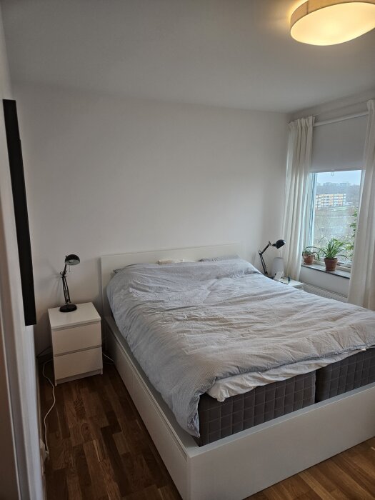 Sovrum med minimal inredning, stor säng och fönster med växter. Diskussion om att hänga tavlor ovanför sängen.