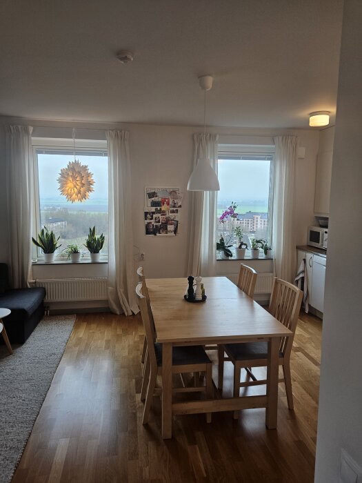 Ljust matsalsutrymme med träbord och stolar, stora fönster med utsikt, vita gardiner, en taklampa och växter på fönsterbrädorna.