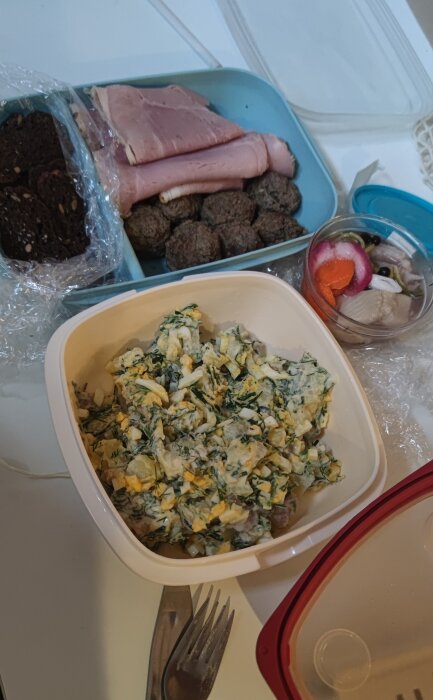 Sill och rotfrukter i burk, potatissallad med ägg och dill, köttbullar, skivad skinka, och rågbröd med frön på en vit yta.