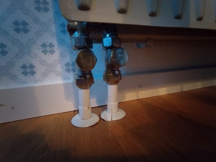 Element med två kopplingar och rör som går ner i ekmönstrat golv; tapet med blommönster i bakgrunden.