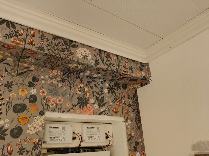 Blommigt tapetserad kökshörna med sned taklist och elbox monterad på väggen.