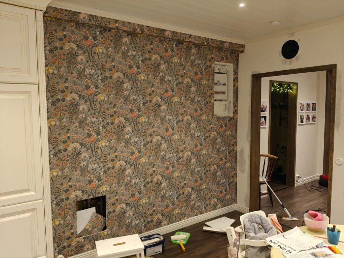En vägg med blommigt tapetmönster, en oklippt del nertill visar mönsterpassning. Verktyg och papper ligger på golvet och bordet.