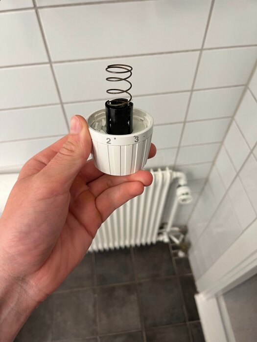 Hand håller trasig termostat med fjäder, vit ratt märkt med siffror, i ett kaklat badrum med radiator i bakgrunden.