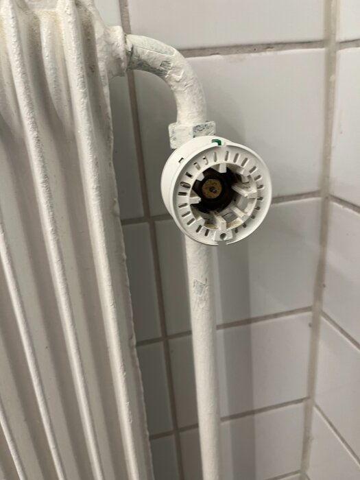 Vit badrumstermostat saknas på en radiator, med kakelvägg i bakgrunden.