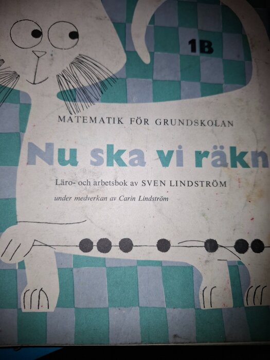 Omslaget till en gammal matematikbok för grundskolan med titeln "Nu ska vi räkna", illustrerat med en tecknad katt mot ett rutigt mönster.