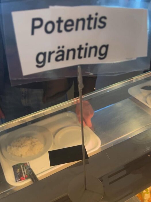 En skylt med texten "Potentis gränting" ovanför en bricka med mat bakom en glasdisk.
