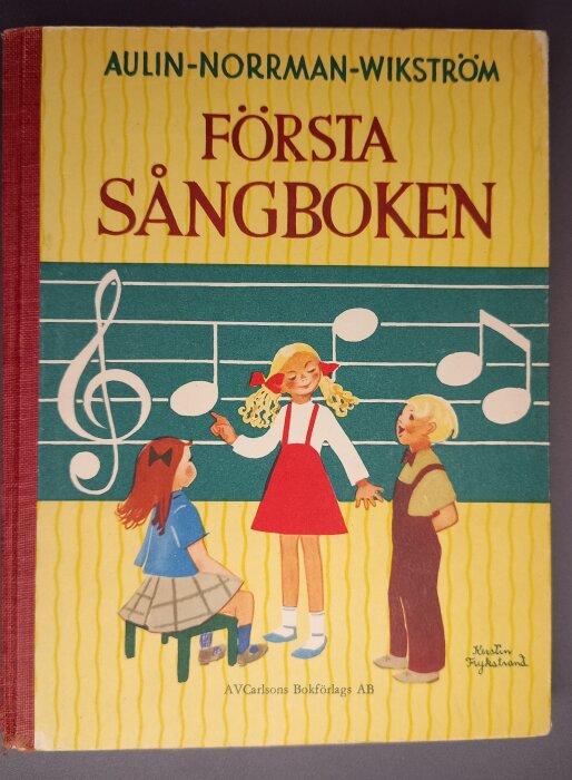 Omslag på sångbok från 1956 med illustration av tre barn och noter, "Första Sångboken" av Aulin-Norrman-Wikström.