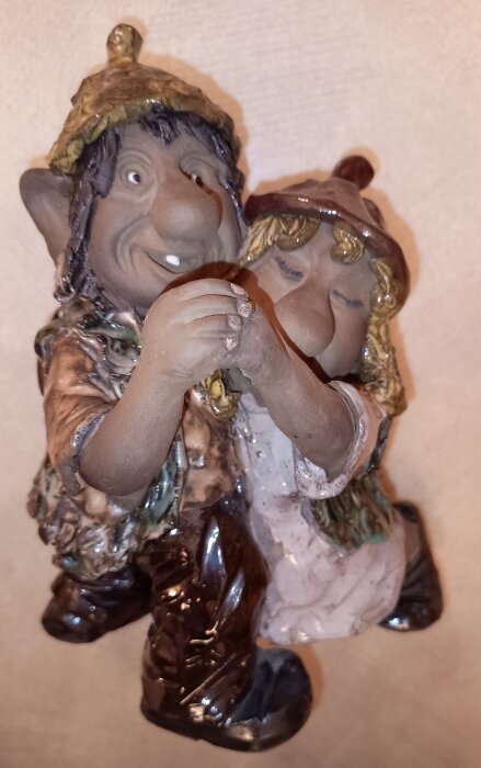 Två keramikfigurer i form av glada troll i färgglada kläder, sittande och handhållande, på en beige bakgrund.