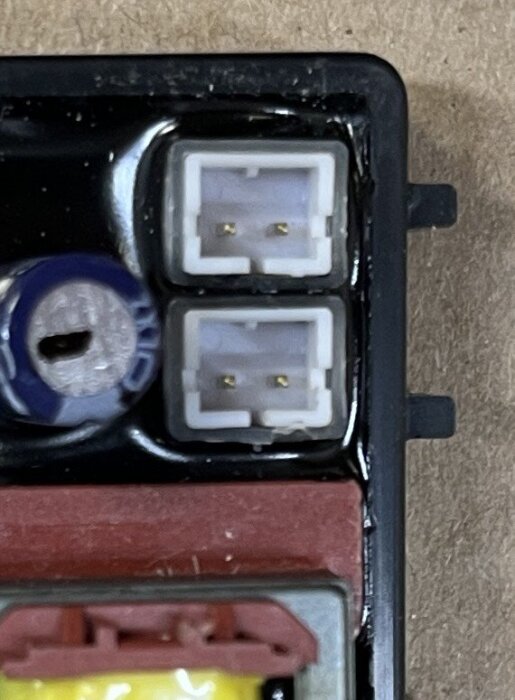 Vit kontakt med två stift i en svart ram, bredvid en blå komponent och ovanför en röd och gul del, används för elektronik.