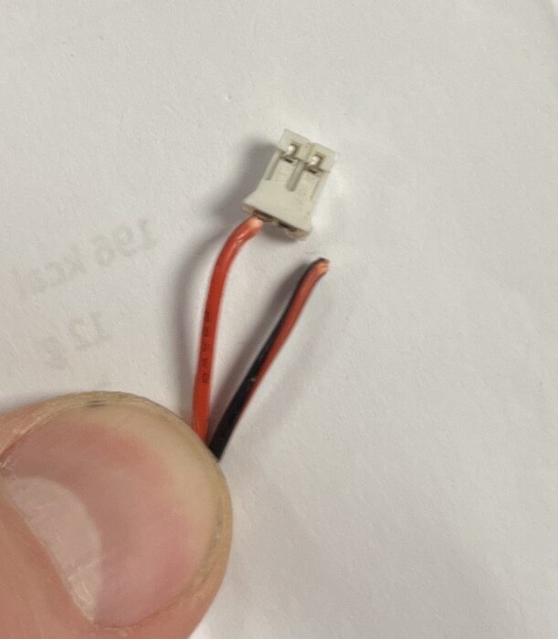 Vit plastkontakt med två ledningar, en röd och en svart, hålls mellan fingrarna på en vit bakgrund.