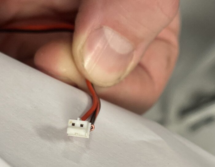 En närbild av en hand som håller en liten vit kontakt med två hål, där röda och svarta kablar är anslutna.