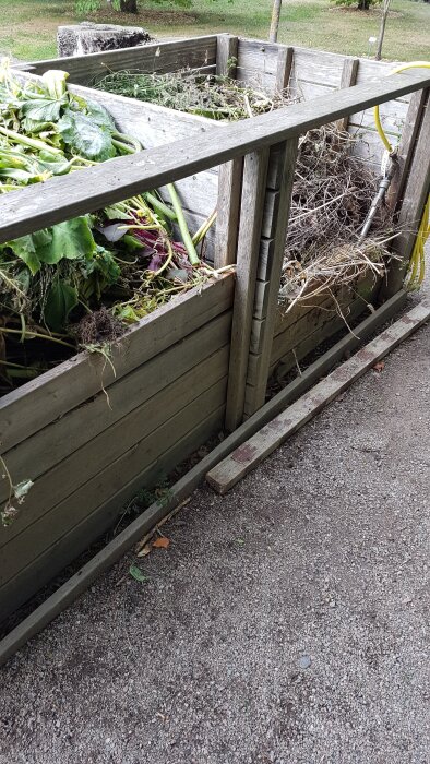 Träkompostlåda med tre fack, fylld med växtrester och placerad på grusmark.