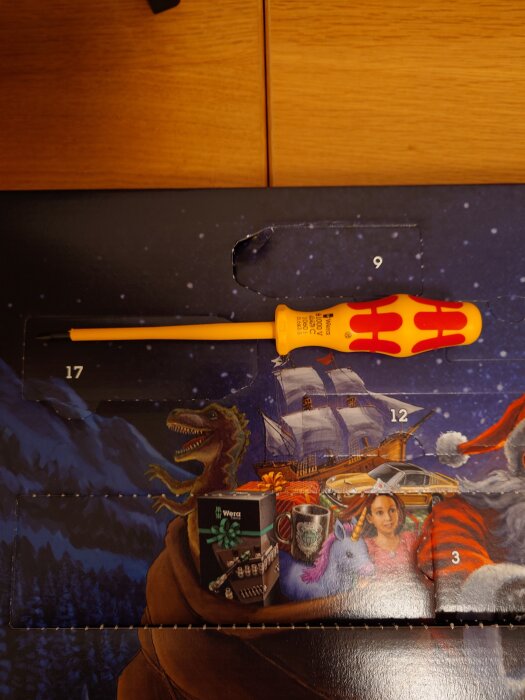 Skruvmejsel med röd och gul handtag placerad över en färgglad bakgrund med en dinosaurie, skepp och julmotiv.