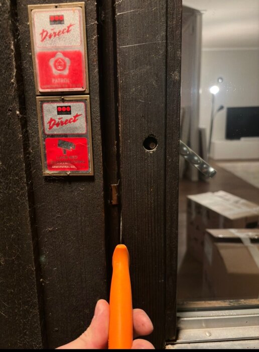 Hand håller orange verktyg som lyfter kilkolv mellan dörr och slutbleck, säkerhetsdekaler synliga på dörrkarmen.