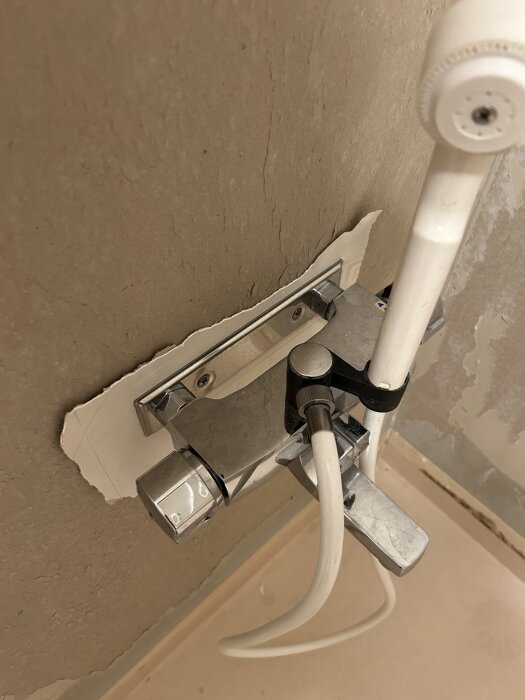 Duschblandare på en rå vägg, med en vit duschslang och duschhandtag. Väggmaterialet ser ofärdigt ut, med delvis borttagen tapet.