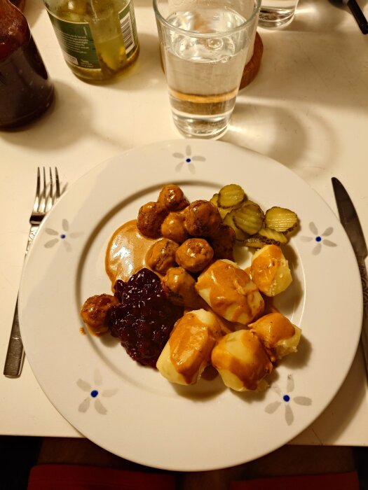 Tallrik med köttbullar, sås, potatis, lingonsylt och skivad gurka, med ett glas vatten och saftflaskor i bakgrunden.