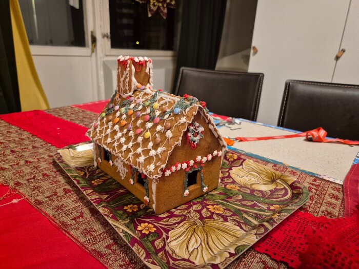 Pepparkakshus dekorerat med färgglatt godis och vit glasyr på ett blomstrande fat, uppställt på ett röd- och guldfärgat bord.