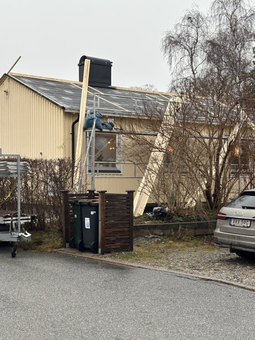 Omläggning av tak på ett hus med byggnadsställningar och brädor, uppställda soptunnor och en parkerad bil i förgrunden.
