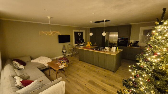 Öppet kök och vardagsrum i julstämning med grön köksö, ljusslingor och julgran i hörnet, beige soffa och träbord, mörkt belysning.