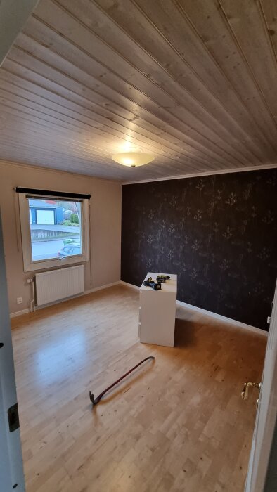 Sovrum under renovering med ljust trägolv, mörk mönstrad vägg och verktyg på låda. Brädpanel i taket och stort fönster med utsikt.