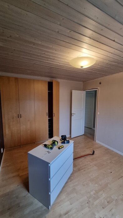 Ett sovrum under renovering med trägolv, vit byrå med verktyg ovanpå, och träpanel i taket. Gula garderober och vit dörr syns också.