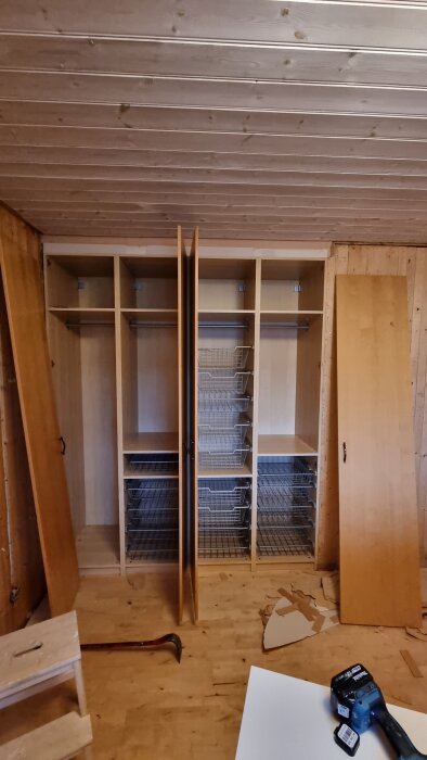 Öppen garderob med trådbackar och klädstänger under montering i rum med trätak och trägolv, omgivna av verktyg och träskivor.