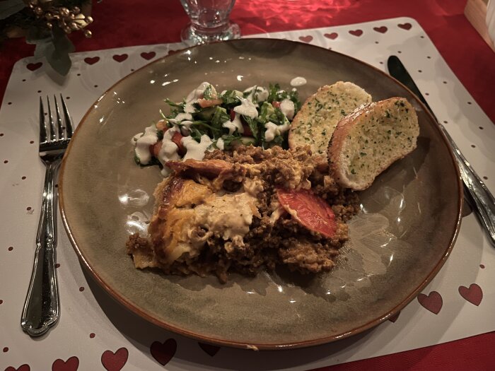 Köttfärspaj med sallad och vitlöksbröd serveras på en tallrik, med hjärtformade dekorationer på duken.