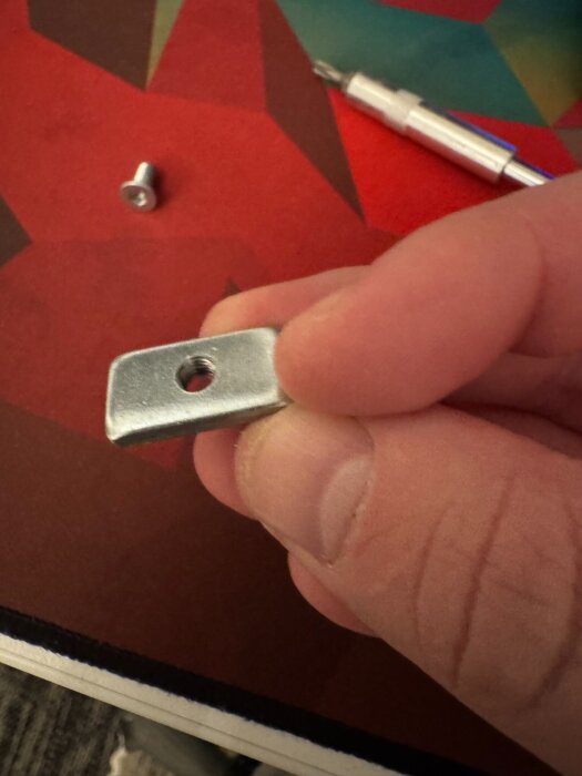 En hand håller en metallbit med ett hål, en liten skruv och ett verktyg ligger i bakgrunden på en färgglad yta.