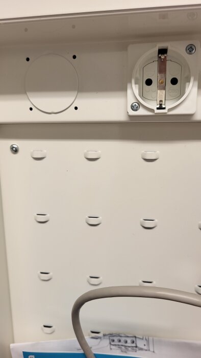 Vit väggpanel med ett eluttag och en kabel framför. En folder med ritningar syns nedtill.