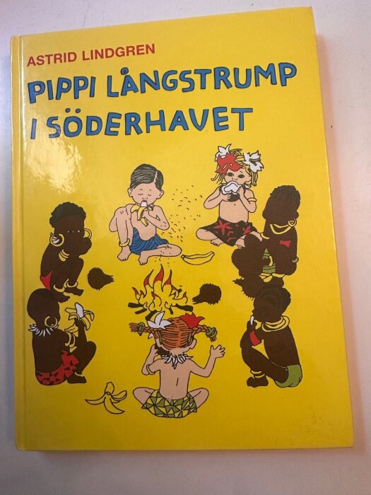 Bokomslag för "Pippi Långstrump i Söderhavet" av Astrid Lindgren, visar Pippi med barn runt en eld, äter bananer.
