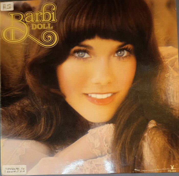 Omslag av LP-skivan "Barbi Doll" från 1974 med en närbild av en leende kvinna med långt brunt hår och ljust klädd i spets.