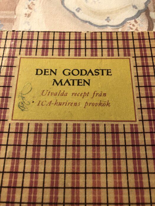 Omslag av 1950-tals kokbok, "Den Godaste Maten", med rutig bakgrund och text från ICA-kurirens provkök.