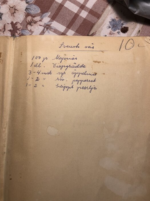 En gammal handskriven receptsida för svensk sås med majonnäs, vispgrädde och kryddor på baksidan av en bok med blomstermönstrad duk i bakgrunden.