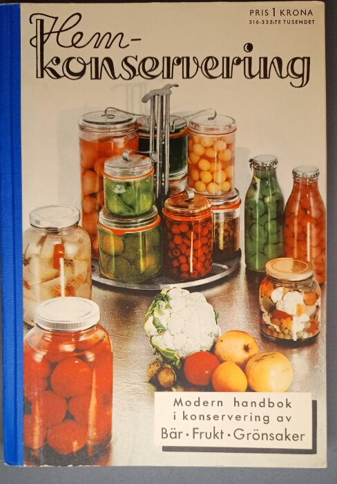 Omslag av bok med titeln "Hemkonservering" med burkar och flaskor fyllda med inlagd frukt och grönsaker, pris: en krona.