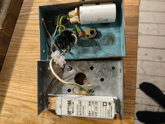 Gammal armaturbox med elektriska komponenter, inklusive en vit kondensator och kablar, på ett träbord.