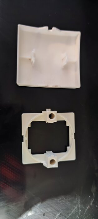 Vit plastdel med fyrkantig ram och cirkulär öppning på svart bakgrund.