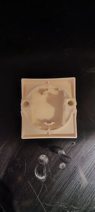 Plastkomponent med två hål och inre detaljer, placerad på en svart yta med repor och märken.