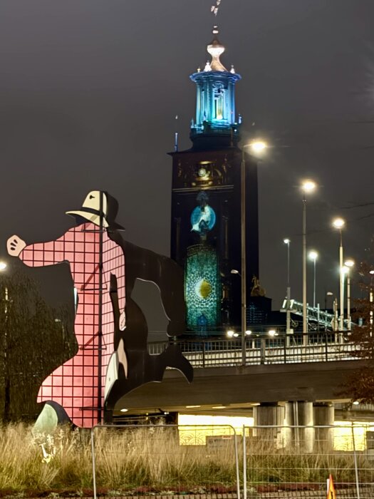 Stor konstinstallation av en figur i hatt framför en stadshusbyggnad med upplyst torn och natthimmel i bakgrunden.