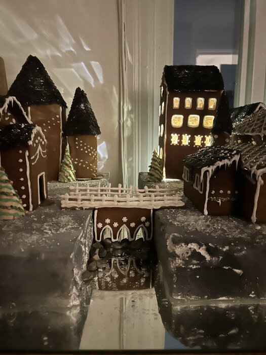 Pepparkakshus med kristyr och belysning, en by med flera byggnader och dekorerade granar, ljus strålar genom fönstren.