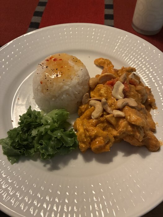 Tallrik med kycklinggryta toppad med cashewnötter, vit portion ris med sås och sallad på en röd bordduk.