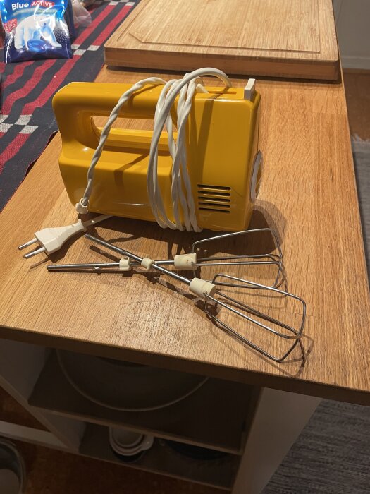 Gul elektrisk handmixer med sladd och vispar placerad på en träbänk i kök.