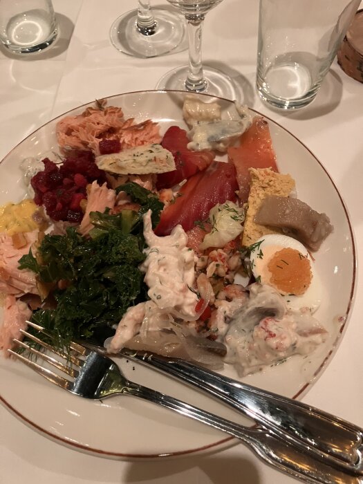Tallrik med olika julbordsmaträtter inklusive lax, rödbetssallad, och grönsaker på vit duk med bestick och glas.