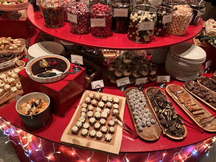 Julbord med olika desserter inklusive chokladpraliner, frukt och bakverk på en röd bordsskiva dekorerad med ljusslingor.