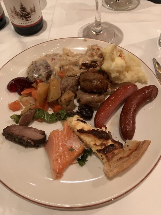 En tallrik med julmatsdelikatesser som sill, korv, lax, kött och potatismos på ett julbord. I bakgrunden en flaska ekologisk öl.