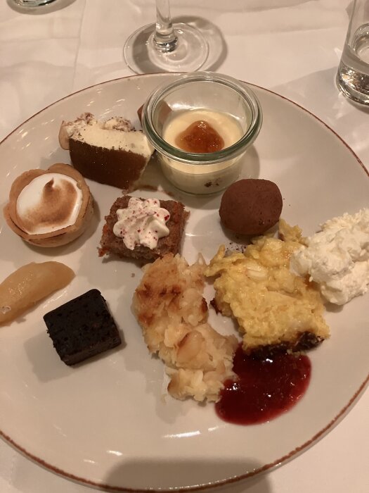 Tallrik med olika julbordets desserter, inklusive mousse, bakverk och fruktpuré, på en vit duk med glas i bakgrunden.