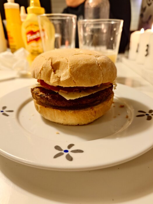 Hamburgare med bröd, ost, och tomat på en tallrik dekorerad med blommönster, omgiven av glas och senapsflaskor.