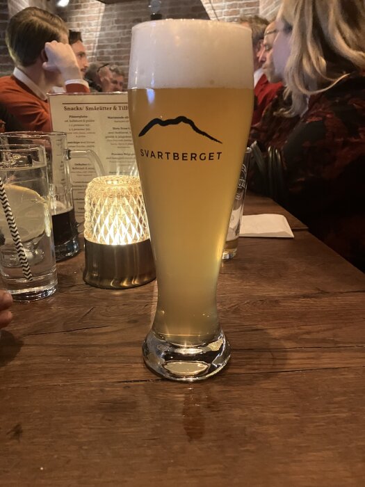 Ölglas med Svartberget-öl på ett träbord, omgivna av en brinnande ljuslykta och sällskap som samtalar.