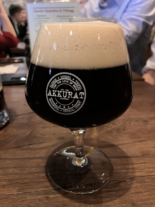 Ett glas mörk öl med skum serverat på ett träbord, märkt med "Akkurat".
