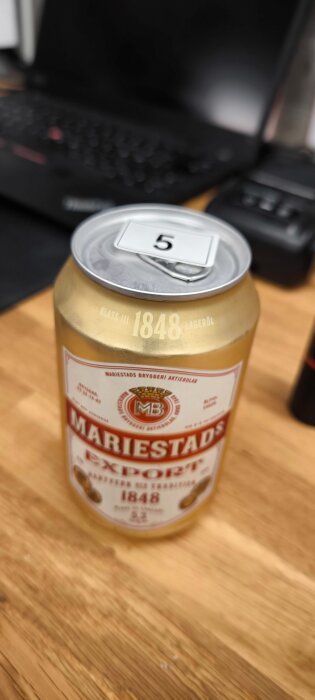 En burk Mariestads Export öl med siffran 5 på locket, står på ett skrivbord vid en laptop.
